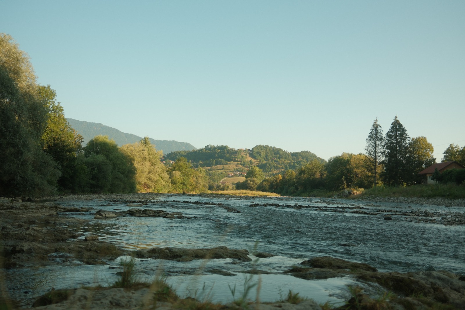Ruhiger Fluss mit Steinen im goldenen Abendlicht, umgeben von grünen Bäumen und Bergen — Sinnbild für die somatische Arbeit und Embodiment Workshops von SOMA Lust in Hamburg